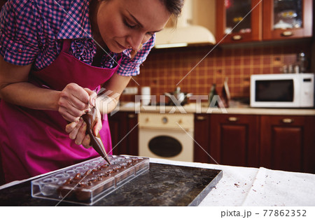 Chef pastry squeezing sweet creamy liquid from confectionery bag into candy molds for making luxury pralines and truffles at home 77862352