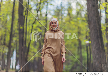 Islamic beautiful woman in a Muslim dress standing on a summer park street background forest autumn trees.world hijab day. 77864376