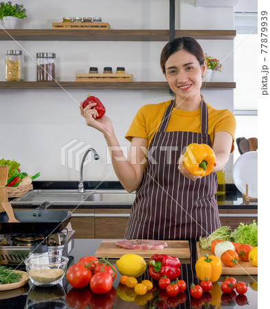 Young housewife stand smiling, hold red and yellow bell pepper with both hands. Offering the yellow one on in the front. The kitchen counter full of various kinds of vegetables. Young housewife stand smiling, hold red and yellow bell pepper with both hands. Offering the yellow one on in the front. The kitchen counter full of various kinds of vegetables. 77879939
