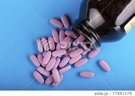 Pink pills are scattered from a glass jar on a blue background.  77891379