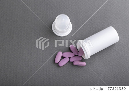 Lilac medical pills are falling out of a white bottle on a gray background. 77891380