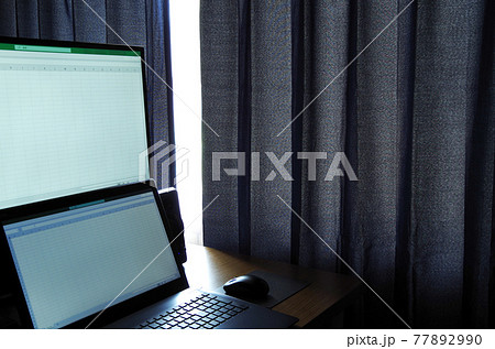 ノートパソコンと増設したモニターの机にカーテンの隙間から屋外の光が差し込んでいる光景 77892990