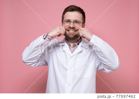 Funny doctor in a white coat makes the artificial smile on face with hands. Stress concept. Pink Funny doctor in a white coat makes the artificial smile on face with hands. Stress concept. Pink 77897462