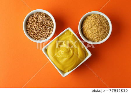 Bowls with mustard seeds, sauce and powder on orange background Bowls with mustard seeds, sauce and powder on orange background 77915359