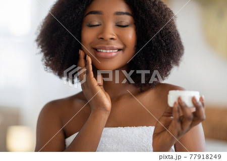 Young African American woman applying hydrating cream on her face, smiling with closed eyes, indoors 77918249