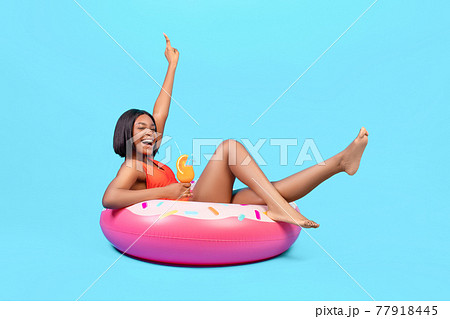 Cool black lady sitting on inflatable ring, having fun, enjoying summer cocktail on blue background, full length 77918445