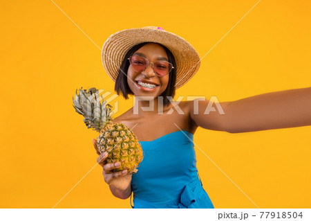 Sexy black lady in trendy summer dress taking selfie with pineapple on orange studio background 77918504