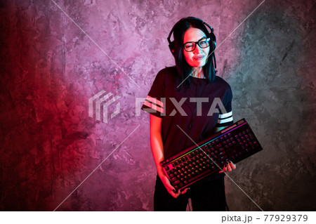 Female esports gamer posing with a gaming gear in neon light. Streamer girl standing with a gaming keyboard. Female esports gamer posing with a gaming gear in neon light. Streamer girl standing with a gaming keyboard. 77929379