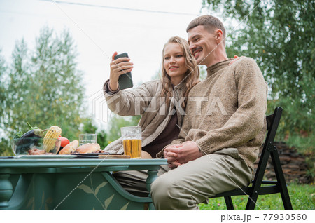 A young couple makes a very funny and emotional selfie, fooling around and making faces in front of the smartphone camera. They made an outdoor dinner with a grill and beer. A young couple makes a very funny and emotional selfie, fooling around and making faces in front of the smartphone camera. They made an outdoor dinner with a grill and beer. 77930560