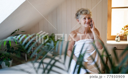 Happy senior woman wrapped in a towel in bathroom at home, resting with tea and smartphone. Happy senior woman wrapped in a towel in bathroom at home, resting with tea and smartphone. 77930909