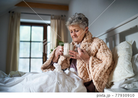 Sick senior woman in bed at home, taking medication. Post-vaccination side-effects. 77930910