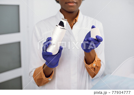 Ultrasonic scrubber device in the hands of a professional female african american doctor beautician in blue gloves. Blurred close up view of young doctor dermatologist posing with ultrasound device. Ultrasonic scrubber device in the hands of a professional female african american doctor beautician in blue gloves. Blurred close up view of young doctor dermatologist posing with ultrasound device. 77934777