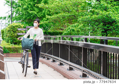 買い物袋を自転車のカゴ（籠）に入れて引いて歩く若い女性  77947634