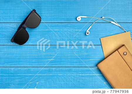 Black sunglasses, white earphones, brown craft paper sheet and leather folder on blue wooden desktop or background. Close up, copy space 77947936