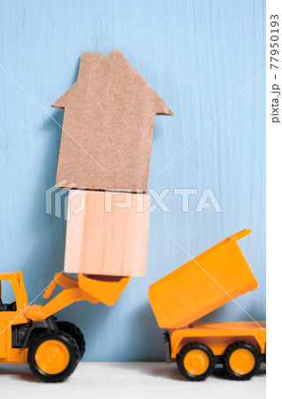 Orange toy excavator loads wooden cube and cardboard house into back of truck. 77950193