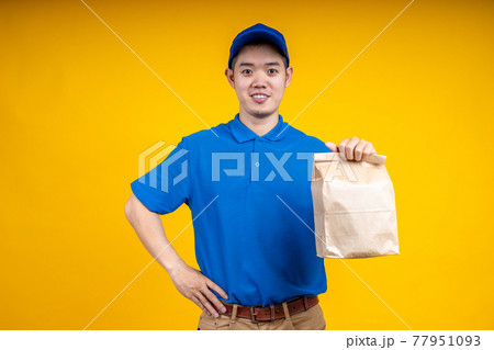 Young Asian handsome delivery man wearing mark holding a food paper bag on right hand over yellow isolate background. Work from home and delivery concept. 77951093