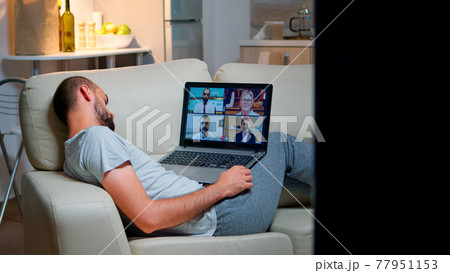 Tired man in pajamas sitting on sofa falling asleep while having online business videocall with teammates Tired man in pajamas sitting on sofa falling asleep while having online business videocall with teammates 77951153
