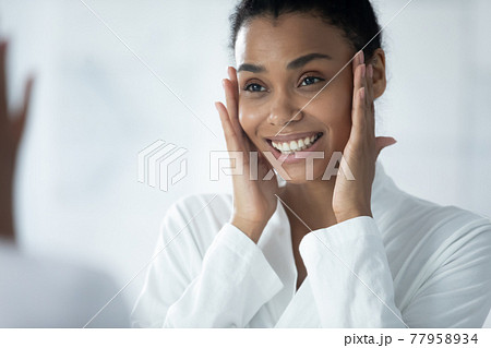 Happy Afro American girl in bathrobe examining smooth facial skin 77958934