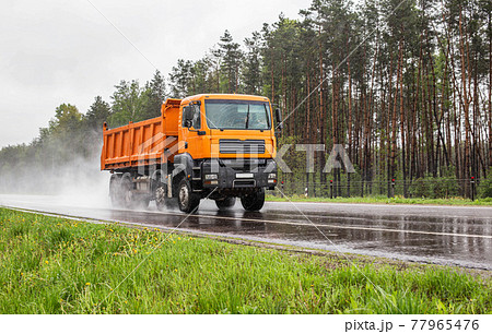 Orange dump truck with 4 axles 40 tonne truck driving on a wet road against the background of a forest, industry. Copy space for text, tonnage Orange dump truck with 4 axles 40 tonne truck driving on a wet road against the background of a forest, industry. Copy space for text, tonnage 77965476