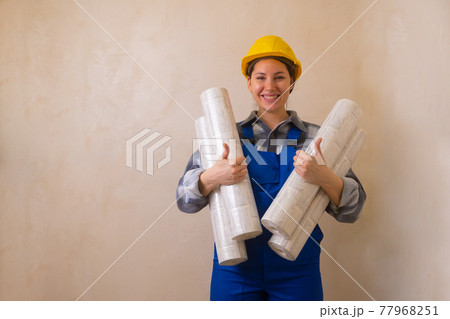 Beautiful brunette construction worker girl holds wallpaper in her hands, smiles and shows thumbs up 77968251