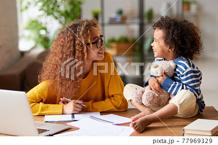 Young happy afro american woman mom talking with little kid son while working on laptop online at home 77969329