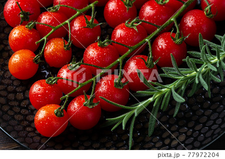 Wet ripe red cherry tomatoes on the vine and sprig of fresh rosemary on a black plate. 77972204