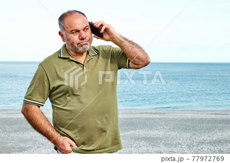 Handsome middle-aged bearded man talking on cell phone while walking on beach with sea in background. Communication and connectivity concept. 77972769