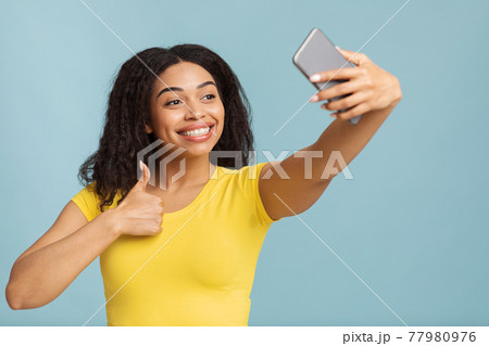 I like it. Happy young black lady taking selfie on smartphone and showing thumb up sign gesture over blue background 77980976