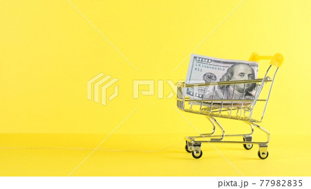 Shopping cart and one hundred dollar bill on a yellow background, closeup. Black Friday Shopping and Shopping cart and one hundred dollar bill on a yellow background, closeup. Black Friday Shopping and 77982835