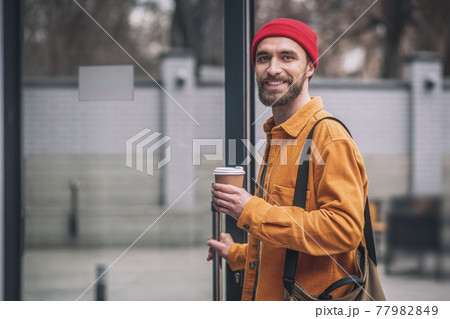 Man in a red hat and orange jacket with a coffee cup in hands looking cheerful 77982849