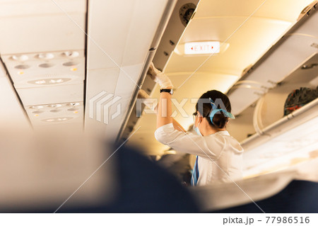 Flight attendant in protective masks and glove closing overhead compartment. Flight attendant in protective masks and glove closing overhead compartment. 77986516