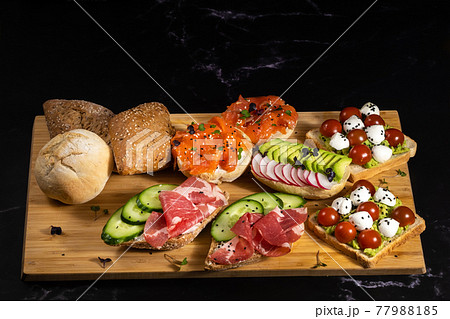 An assortment of sandwiches with fish, cheese, meat and vegetables lay on the board and a bun 77988185