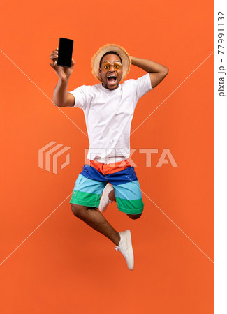 Excited young black guy jumping on air, showing cellphone with black empty screen on orange background, full length 77991132