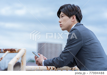 曇り空の屋上でスマホを手にたそがれる若いビジネスマン 77994488