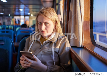 Ferry passenger cabin, sad woman texting with someone online using smartphone. 77998730