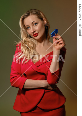 Blonde female in red dress and black earrings. Smiling, showing two blue chips, posing on colorful background. Poker, casino. Close-up Blonde female in red dress and black earrings. Smiling, showing two blue chips, posing on colorful background. Poker, casino. Close-up 78014494