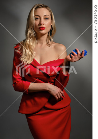 Blonde lady with bare shoulder, in red dress and black earrings. She showing four blue chips, posing on gray background. Poker, casino. Close-up Blonde lady with bare shoulder, in red dress and black earrings. She showing four blue chips, posing on gray background. Poker, casino. Close-up 78014505