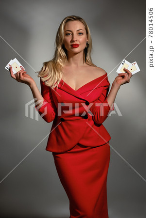 Blonde model in red dress and black earrings. She is showing four aces, posing on gray studio background. Poker, casino. Close-up, copy space 78014506