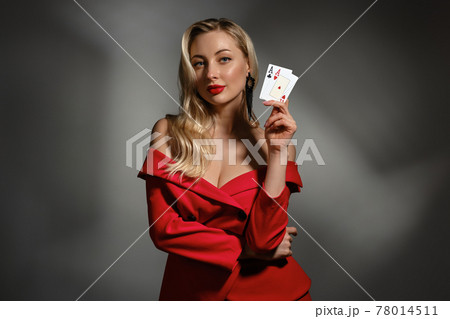 Blonde model in red dress and black earrings. She is showing two aces, posing on gray studio background. Poker, casino. Close-up, copy space 78014511