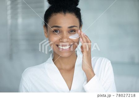 Happy woman in bathrobe applying mask for under-eye skin treatment Happy woman in bathrobe applying mask for under-eye skin treatment 78027218