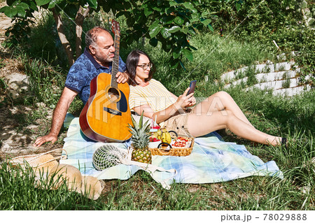 Middle aged couple having a picnic in the garden with fresh exotic fruit and sweet sandwiches. Man in blue t-shirt with floral print near woman makes a selfie 78029888
