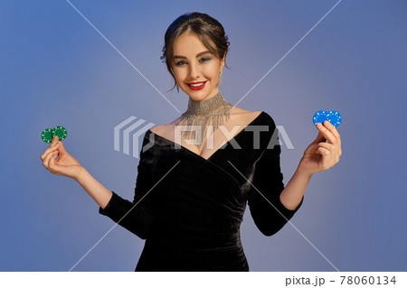 Brunette lady in black dress and shiny jewelry. Smiling, showing two blue and green chips, posing on purple background. Poker, casino. Close-up Brunette lady in black dress and shiny jewelry. Smiling, showing two blue and green chips, posing on purple background. Poker, casino. Close-up 78060134