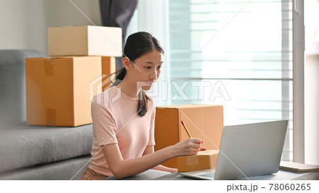 Beautiful girl writing costumer address on box while sitting at the modern living room. SME business startup. Beautiful girl writing costumer address on box while sitting at the modern living room. SME business startup. 78060265