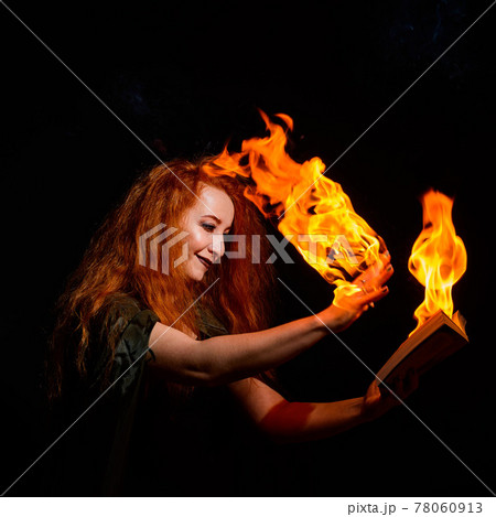 An ominous witch sets her spell book on fire with the power of thought. Red-haired woman conjures for Halloween. Flames on the hands. 78060913