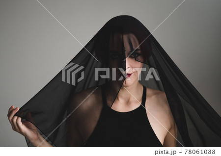 Redhead woman with a black veil on her head in a dark studio. A girl with an earring in her nose smiles slyly. Witch Makeup. Halloween costume. 78061088