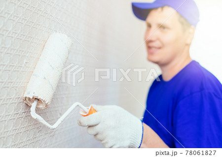 A painter in a cap and gloves paints the wall with a paint roller. Portrait of a young worker in 78061827