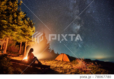 Female traveler sitting near campfire under night starry sky. 78067780