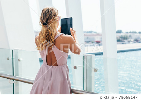 A young woman in a summer dress takes a photo of the seascape on a tablet while walking around the city A young woman in a summer dress takes a photo of the seascape on a tablet while walking around the city 78068147