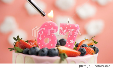 Birthday cake number 67, pink candle on beautiful cake with berries and lighter with fire against background of white clouds and pink sky. Close-up 78089302