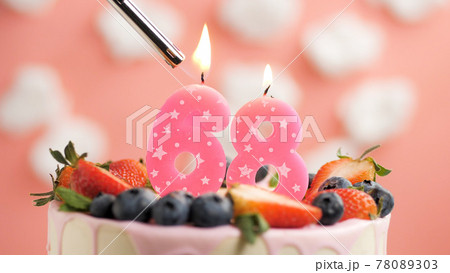 Birthday cake number 68, pink candle on beautiful cake with berries and lighter with fire against background of white clouds and pink sky. Close-up 78089303